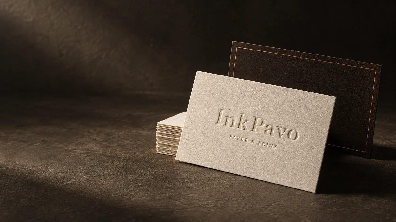 Concept image of an ivory letterpress card reading Inkpavo, Paper & Print, standing against a dark card