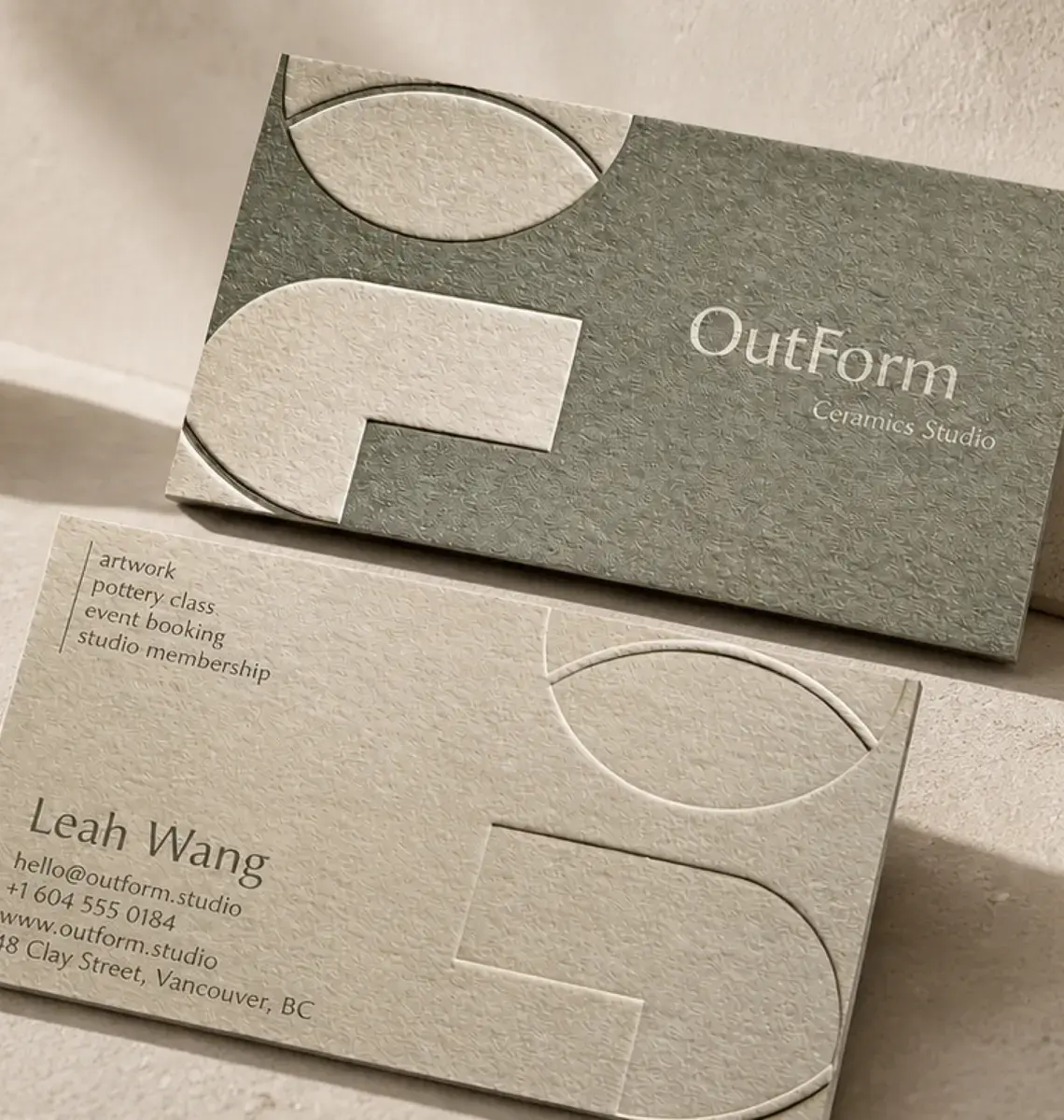 A sage green and a cream uncoated business card for a ceramics studio, with blind-debossed shapes, on a sunlit table