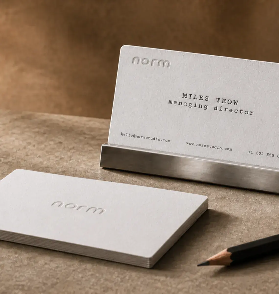 Two grey cotton letterpress business cards, one standing in a steel holder, beside a pencil