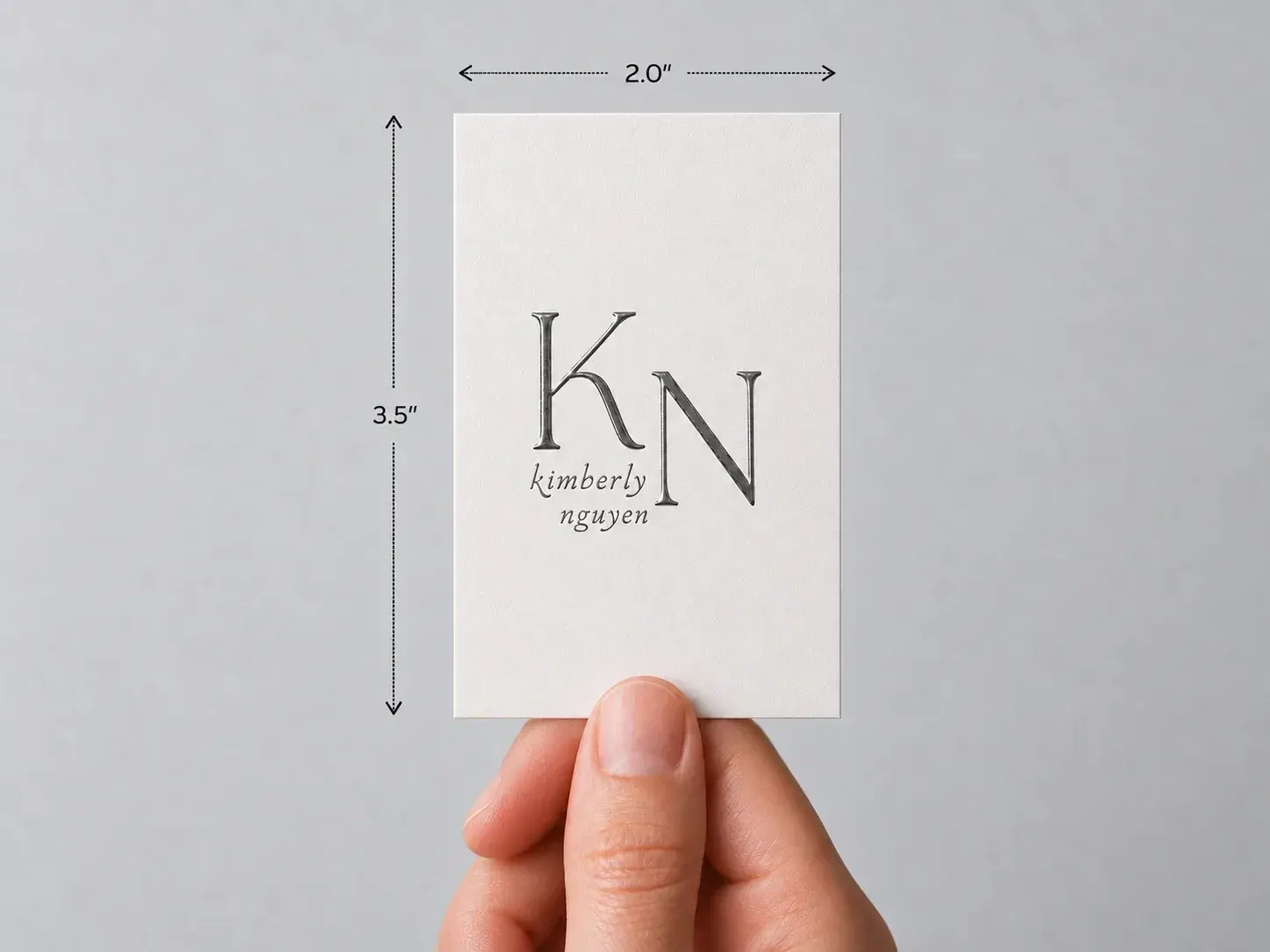 A hand holding an upright white card with a large monogram in raised UV, marked 2 × 3.5 in