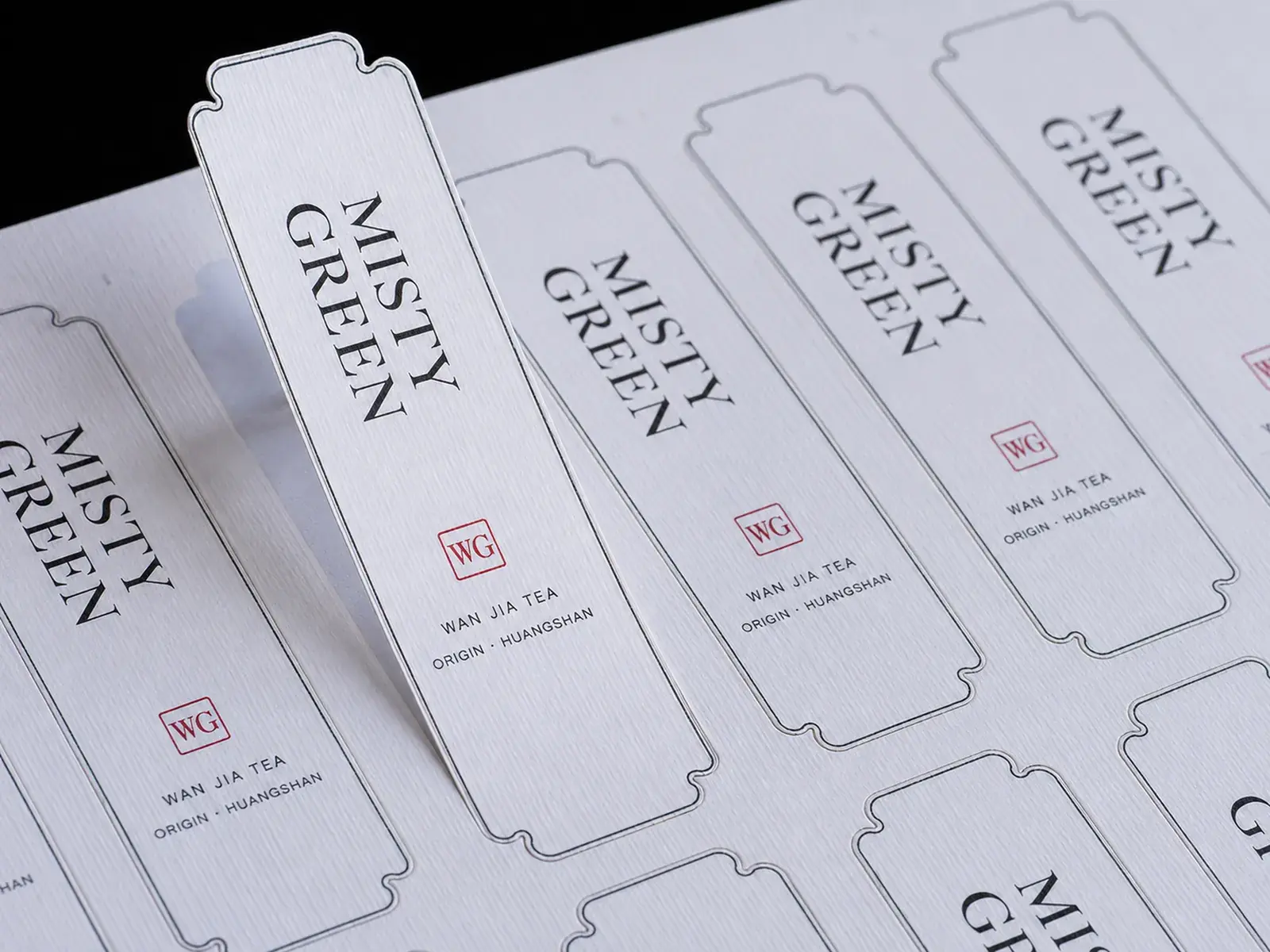 A sheet of ticket-shaped tea labels on white laid paper, one lifted to show the kiss-cut outline