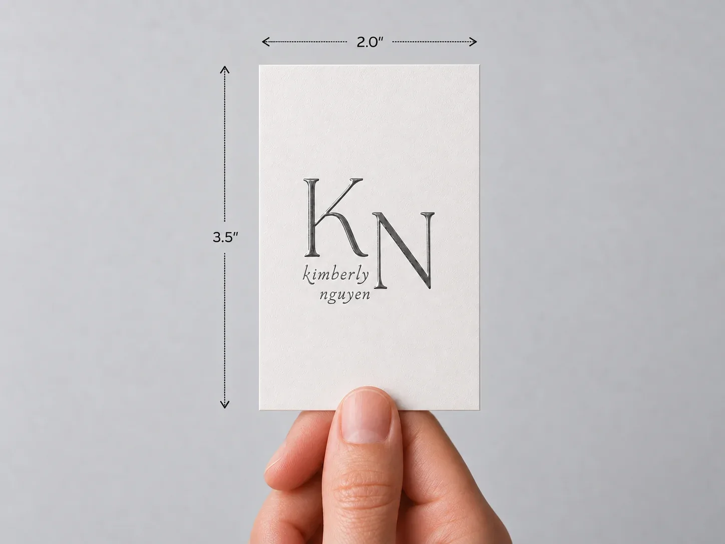 A hand holding an upright white card with a large monogram in raised UV, marked 2 × 3.5 in