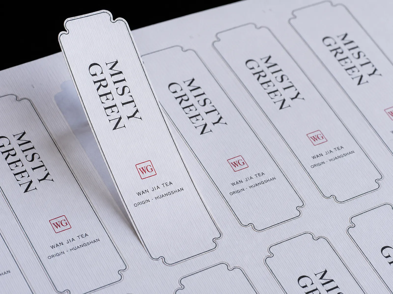A sheet of ticket-shaped tea labels on white laid paper, one lifted to show the kiss-cut outline